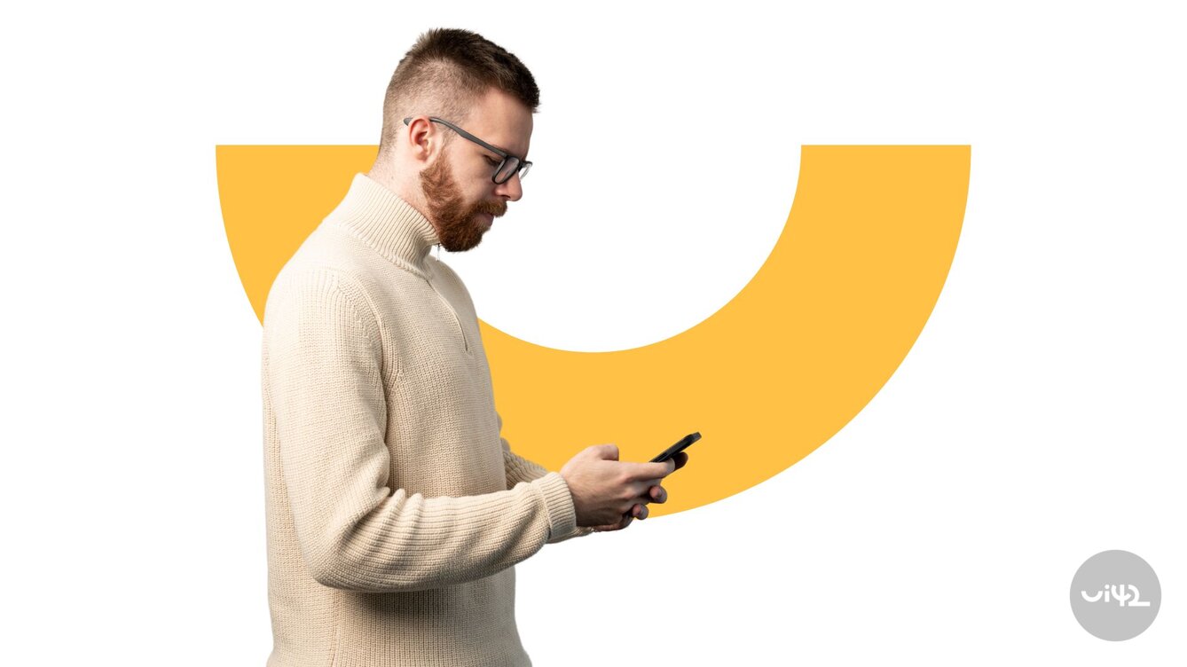 The man in the beige sweater is holding a mobile phone, with a yellow graphic element in the background.