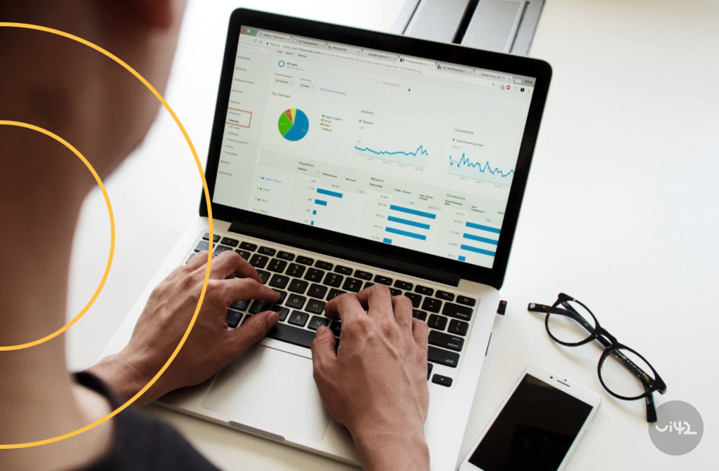 A person working on a laptop with statistics displayed on the screen, next to it are glasses and a mobile phone.